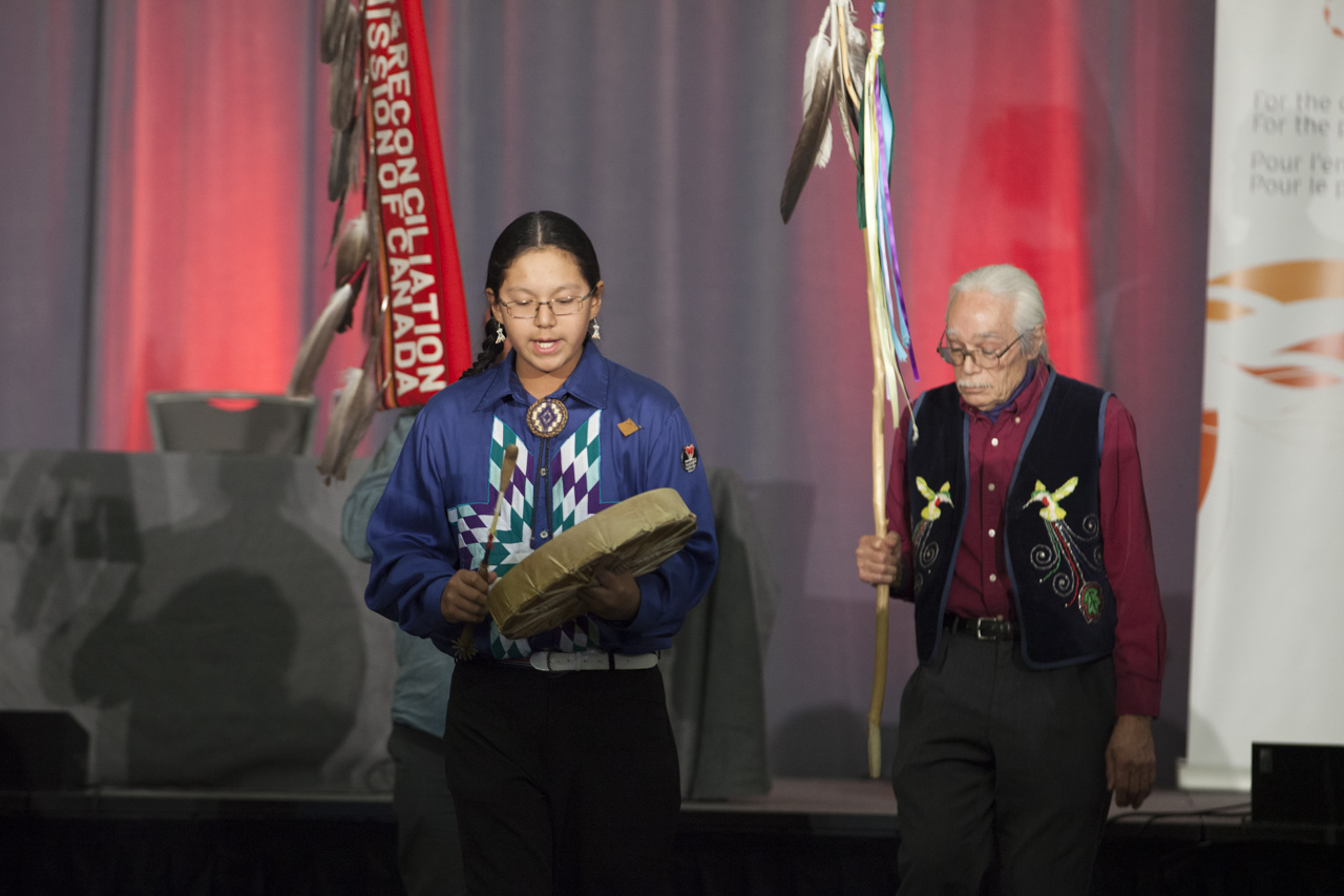 Young man drumming at TRC closing ceremony with Elder standing to his left holding a feather and ribbon adorned staff