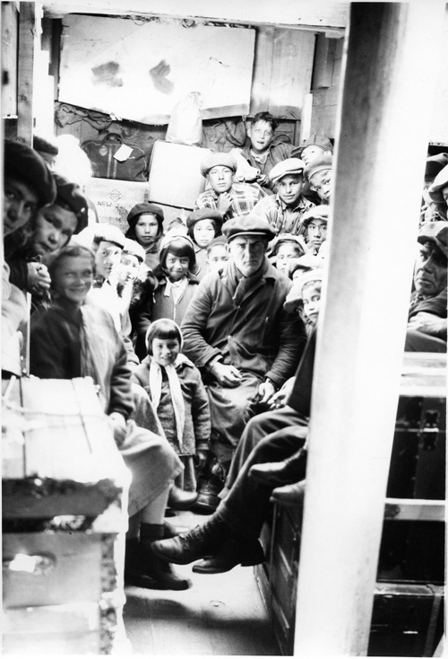 Image en noir et blanc d’un homme blanc renfrogné entouré de plus d’une vingtaine d’enfants autochtones dans ce qui pourrait être un wagon de chemin de fer.