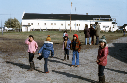 Photo couleur des années 1950 montrant des enfants autochtones debout sur un chemin et dans un champ, vêtus de vêtements d’hiver, avec un long bâtiment blanc en arrière-plan.