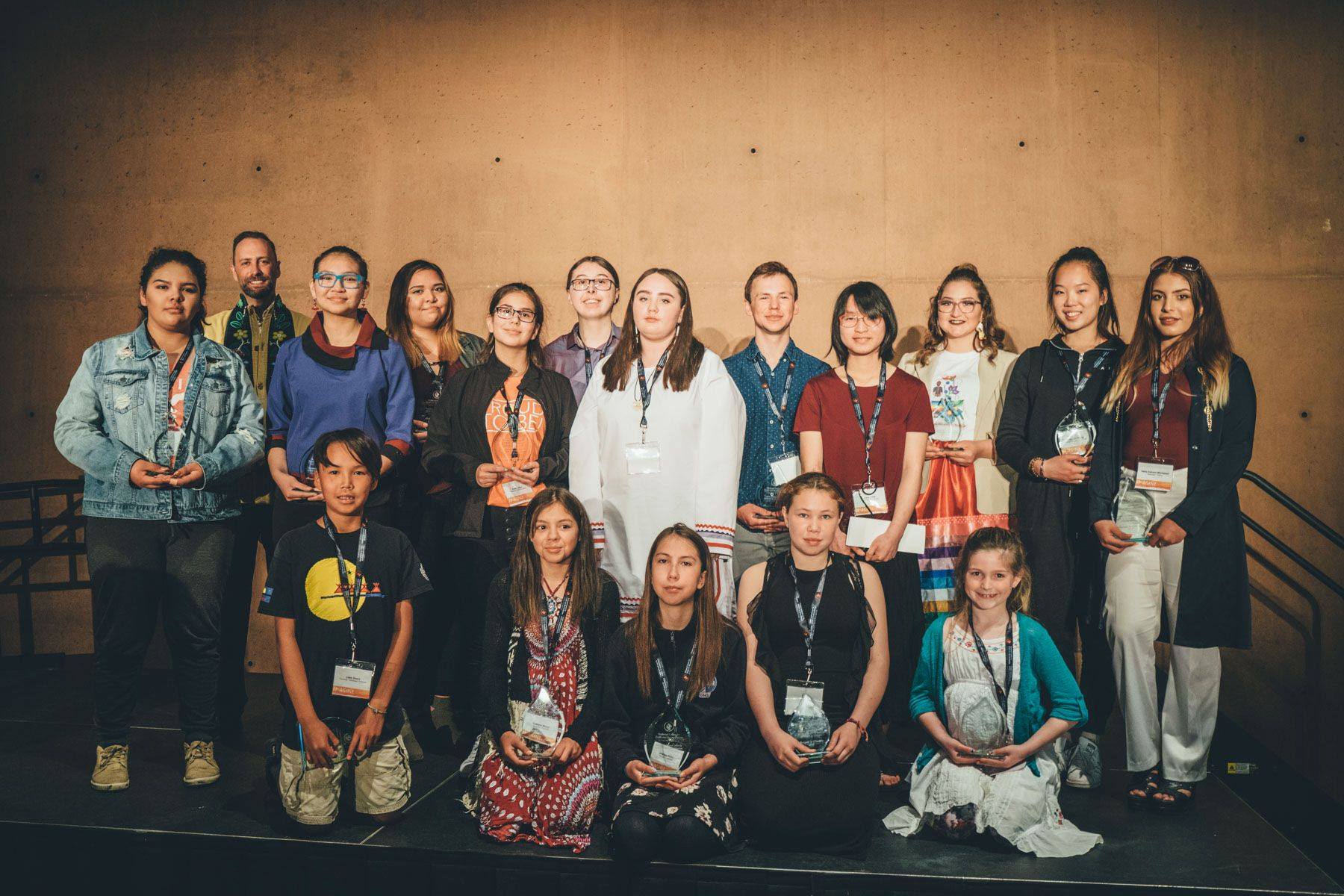 Un groupe de 17 étudiants et enseignants posent devant une toile de fond de couleur brun clair lors d'un événement Imaginez le Canada.