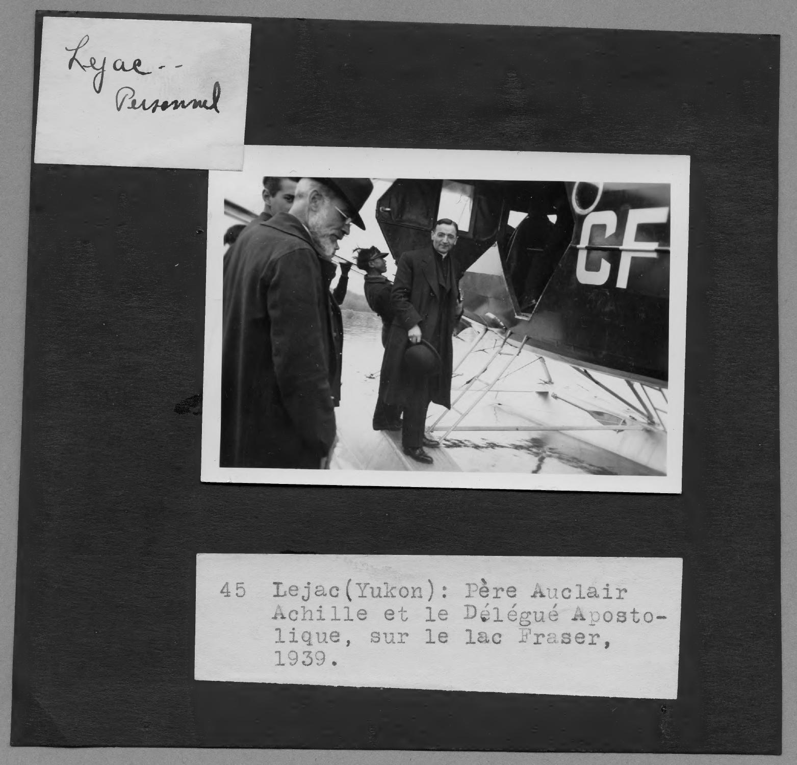 A black and white photograph laid on a black background. In the photo, a priest and three other men stand near a small plane. Below the photo, there is a text that reads “Père Auclair Achille et le Délégué Apostolique, sur le lac Fraser, 1939.”
