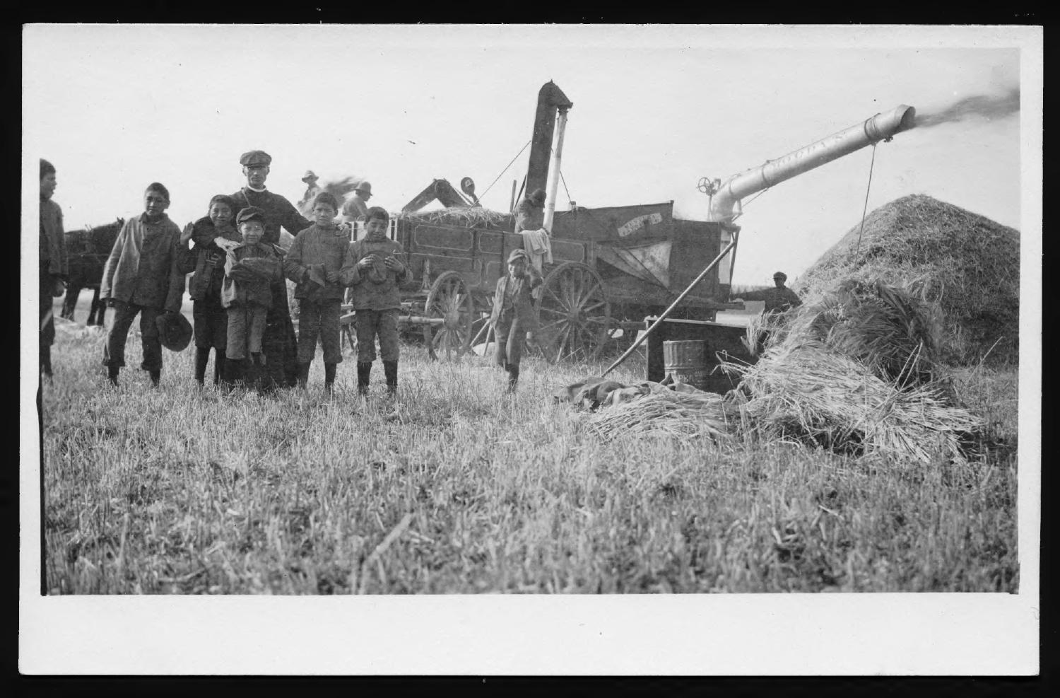 Father Eugène Baillargeon stands with a group of children posing in front of farming equipment.
