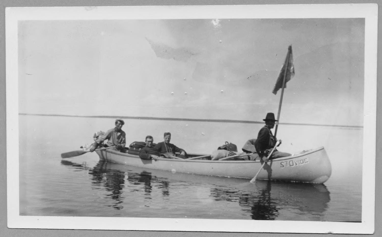 Four men sit in a canoe called the St. Ovide. They are in the middle of a large lake.
