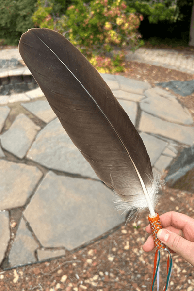 A hand holds a large eagle feather with an orange and silver beaded handle and colourful ribbons.