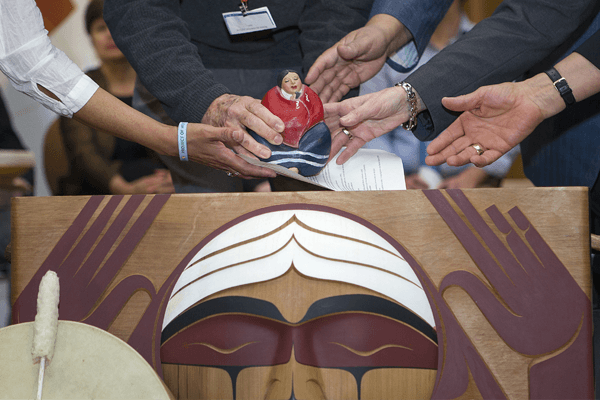 Multiple hands place a small figurine into the Bentwood Box.