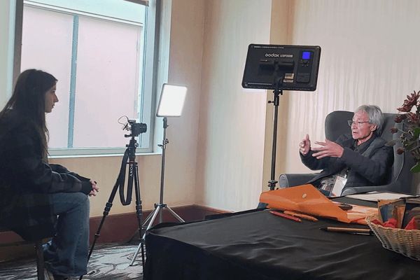 An older person is interviewed by a young woman in a room with lighting and a camera.